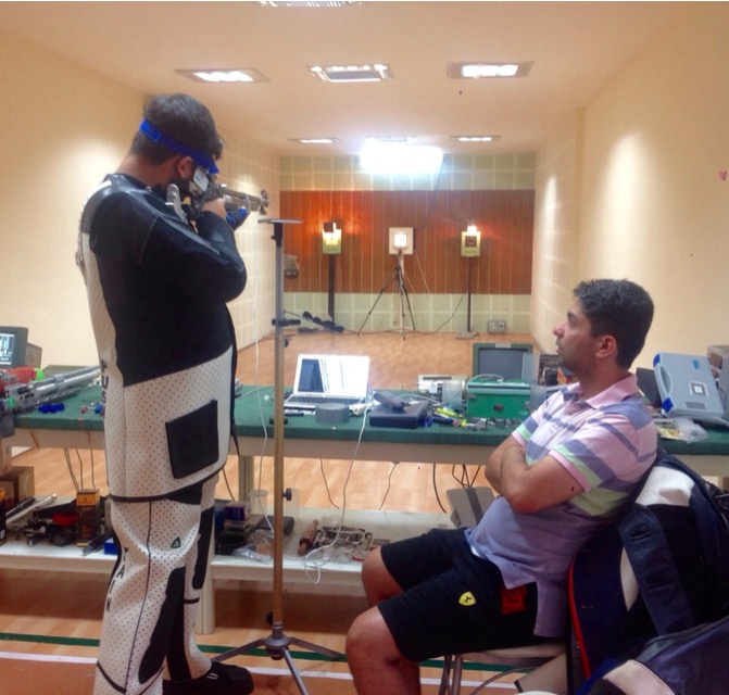 A shooter in standing position aims an air rifle at electronic targets in an indoor range, while a seated Abhinav Bindra watches