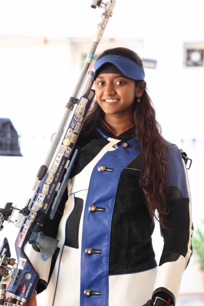 Elavenil Valarivan, wearing her shooting jacket and cap, smiling with her air rifle over her shoulder